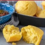 A split corn muffin sits in front of a muffin tin filled with fresh corn muffins; a blue bowl of chili is partially visible on the left.