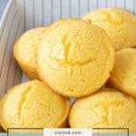 A tray of golden corn muffins is arranged in a stack on a striped surface, showcasing the delicious texture of these classic corn muffins, with a gray cloth in the background.