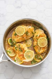 Skillet with cooked chicken breasts, lemon, and parsley.