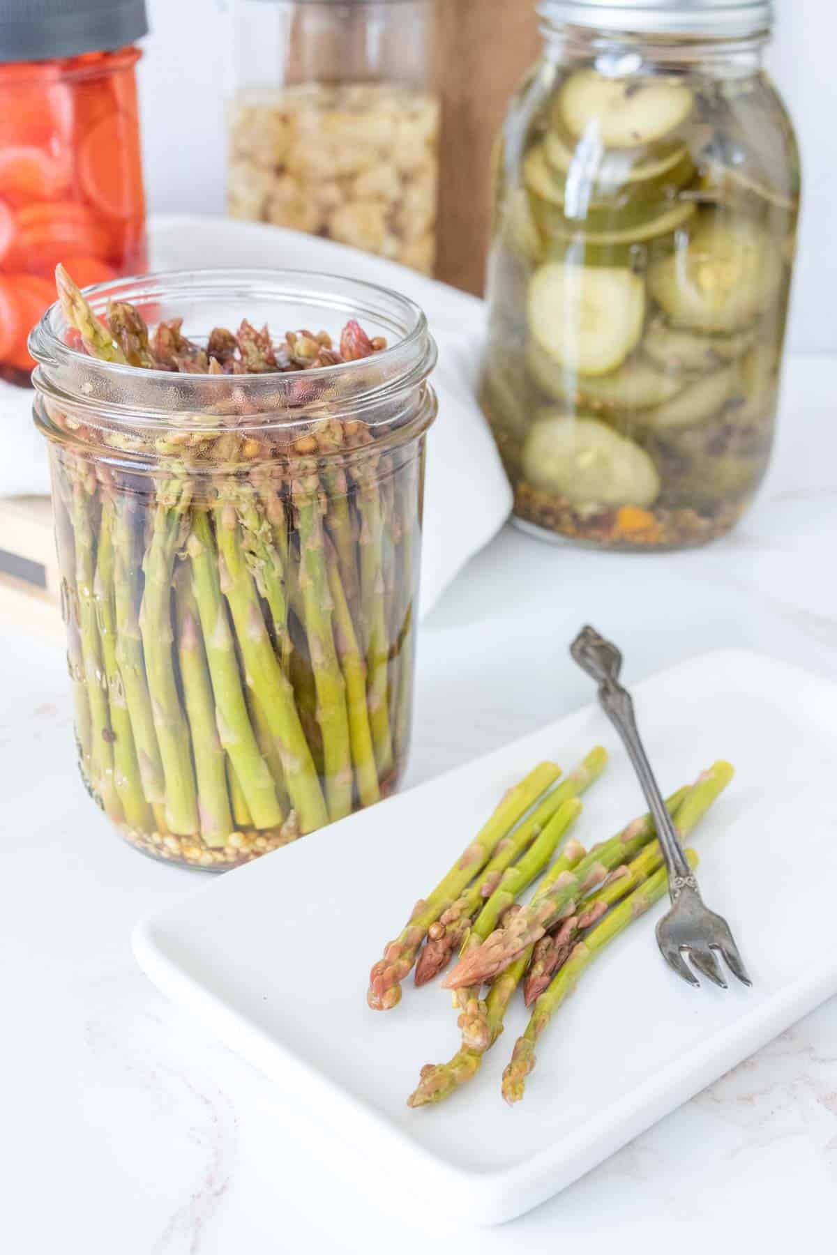 Jar of pickled asparagus with some pickles on a plate and more pickle jars behind.