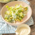 Caesar salad dressing in a ceramic cruet with a Caesar salad behind.