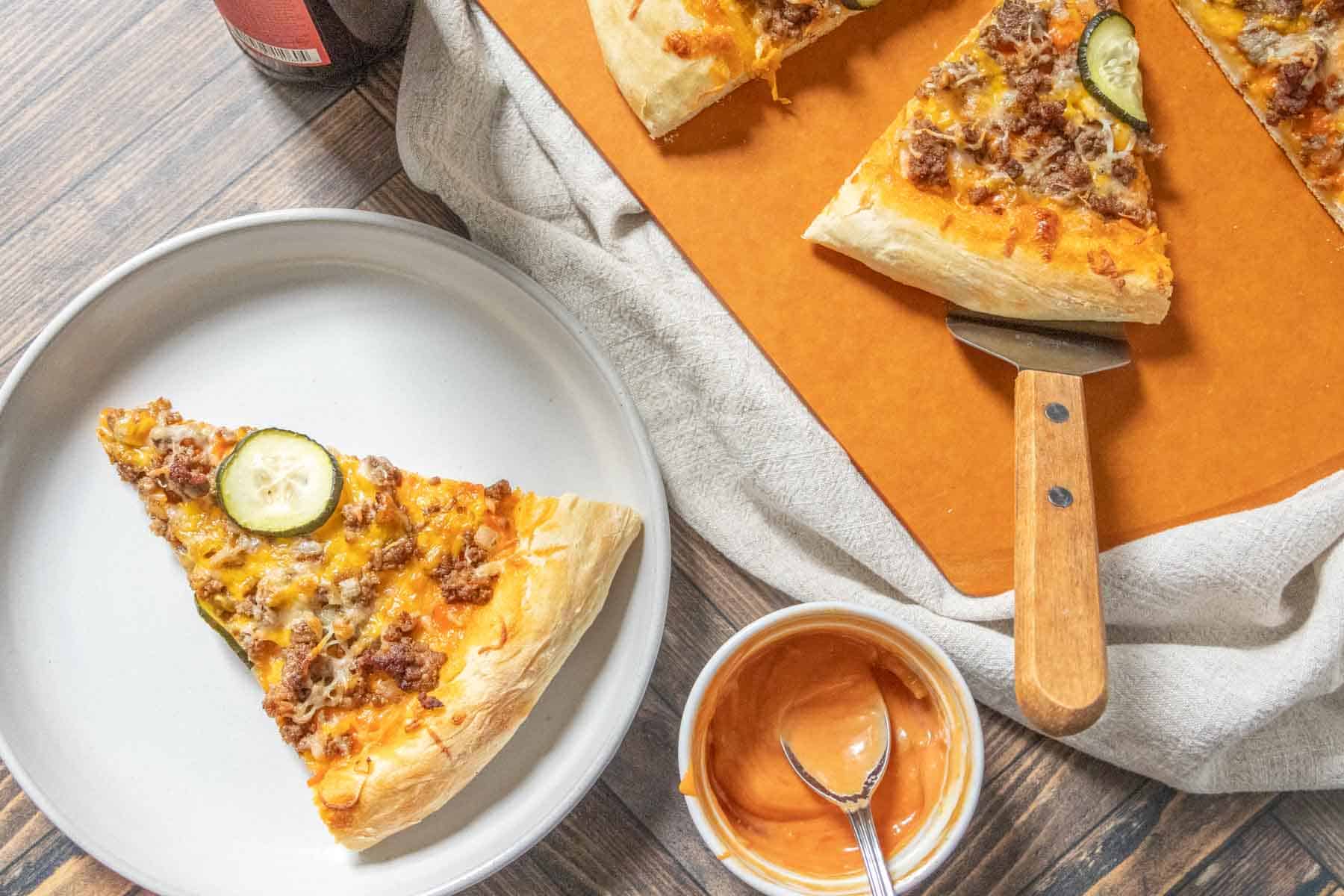 Overhead of a slice of cheeseburger pizza on a plate with the rest of the pizza beside.