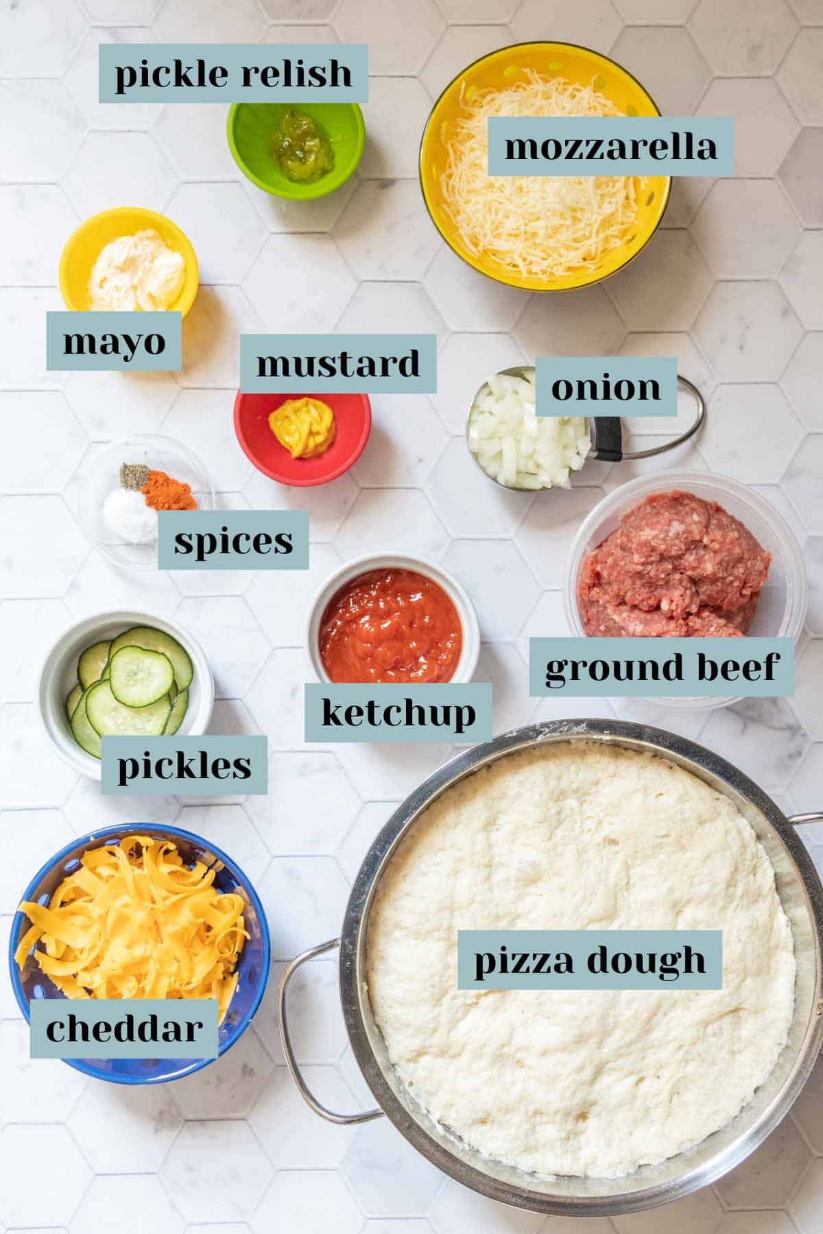 Ingredients for cheeseburger pizza on a tile surface with labels.