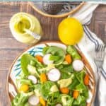 A plate of fresh salad with spinach, carrots, radishes, and avocado sits next to a jar of creamy avocado dressing, a whole avocado, and a lemon on a wooden table. Perfect for those who love vibrant flavors and creamy avocado dressing.