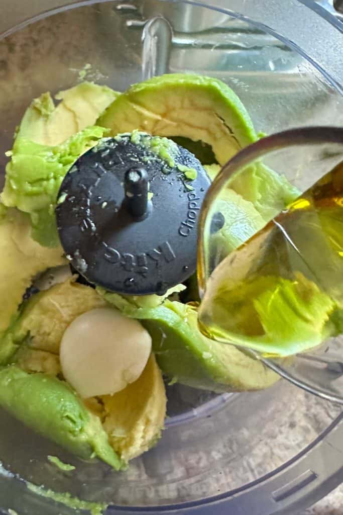 Adding olive oil to a food processor with avocado and garlic.