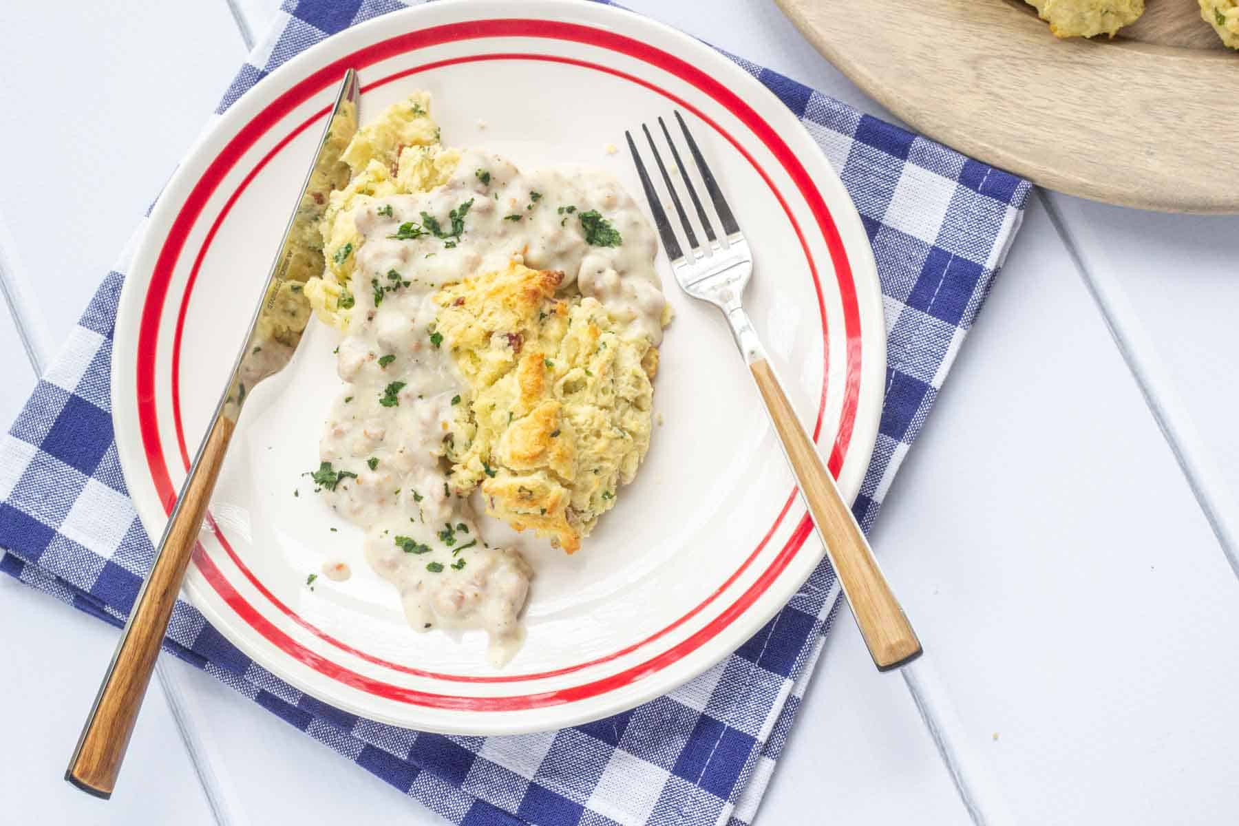 Overhead of a white plate with red rim on a blue checked napkin, with a biscuit and gravy on the plate along with a fork and knife.