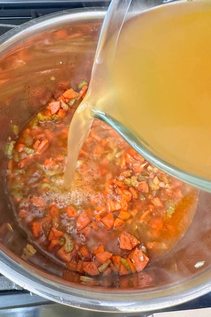 Pouring broth into a soup pot with vegetables in it.