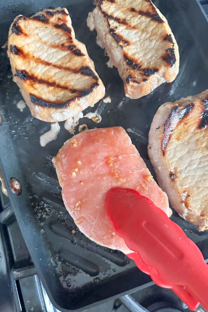 Turning pork chops cooking on a grill pan with red tongs.