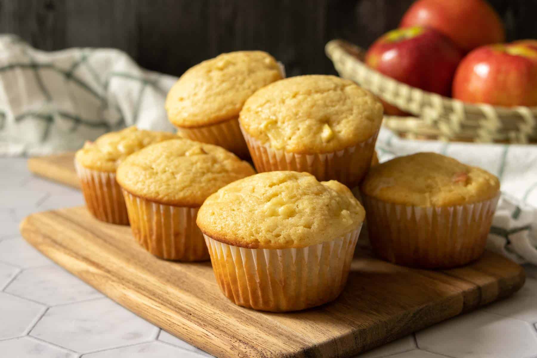 Maple apple muffins on a cutting board with a basket of apples behind.