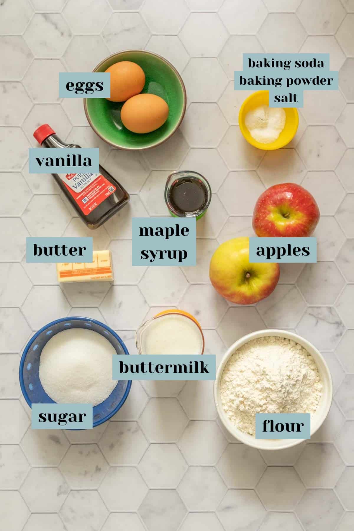 Ingredients for apple muffins on a tile surface with labels.
