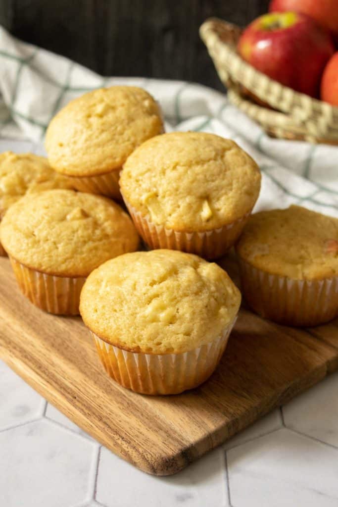 Apple muffins stacked on a wooden cutting board with bowl of apples behind.