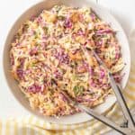 Homemade coleslaw in a serving bowl on a white wooden surface with a yellow striped kitchen towel and tongs.