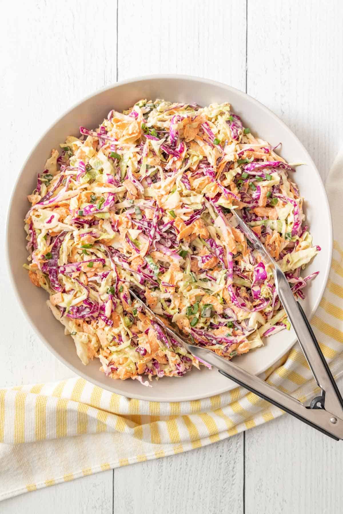 Homemade coleslaw in a serving bowl on a white wooden surface with a yellow striped kitchen towel and tongs.