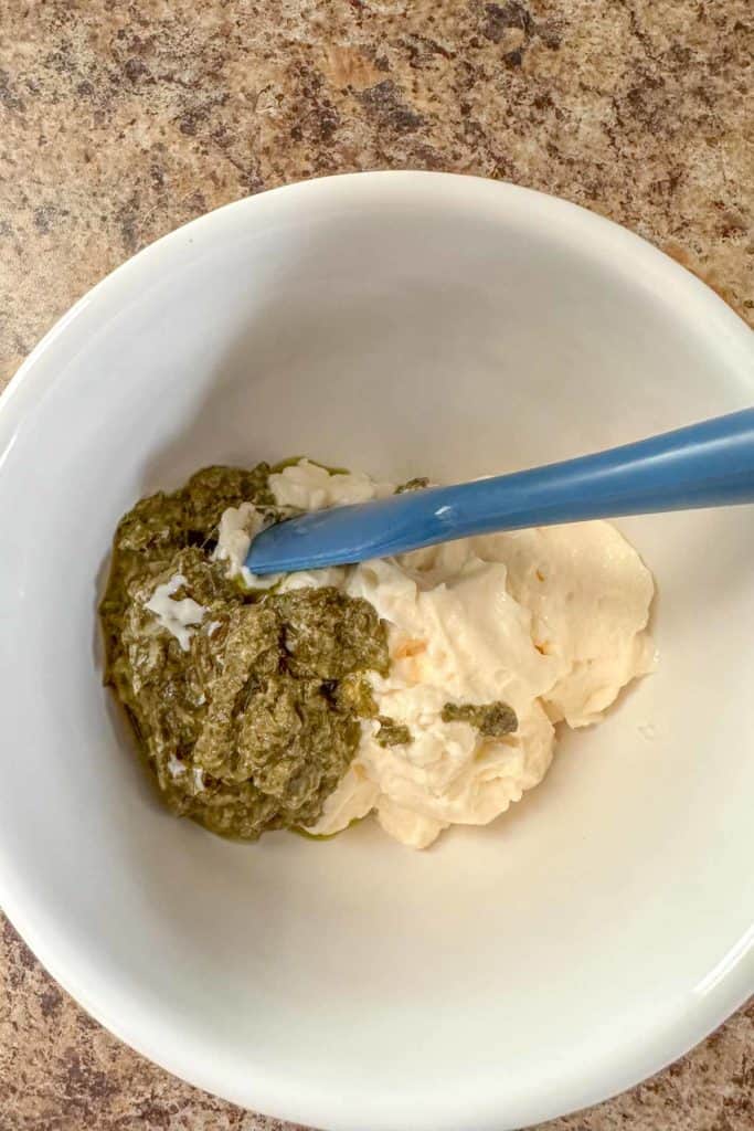 Mayonnaise and pesto in a white bowl before mixing together.