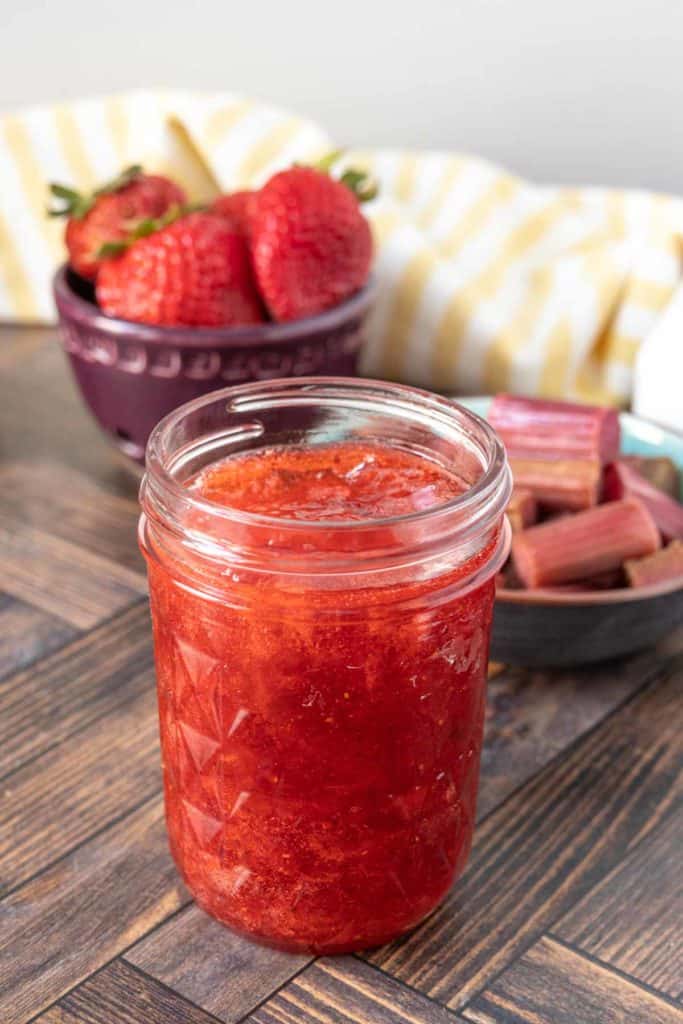 An open jar of strawberry rhubarb jam in front of bowls of strawberries and rhubarb.