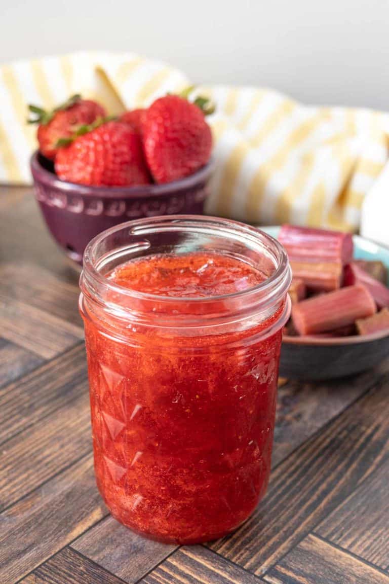 An open jar of strawberry rhubarb jam in front of bowls of strawberries and rhubarb.