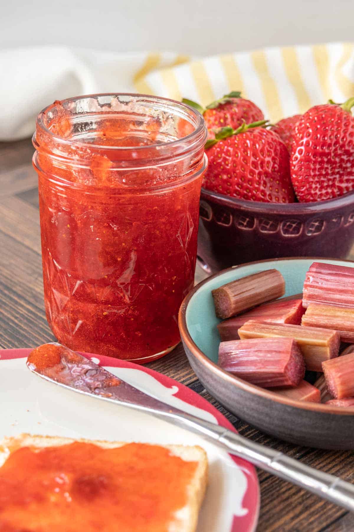 Open jar of strawberry rhubarb jam alongside bowls of strawberries and rhubarb, with a plate of toast in the front.