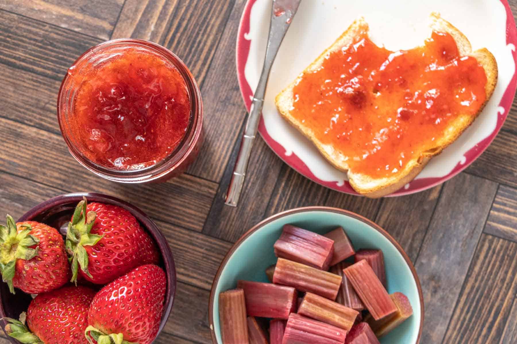 Overhead of strawberry rhubarb jam in a jar next to bowls of strawberries and rhubarb.