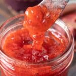 A spreader holding up a dollop of strawberry rhubarb jam just above the jam jar.