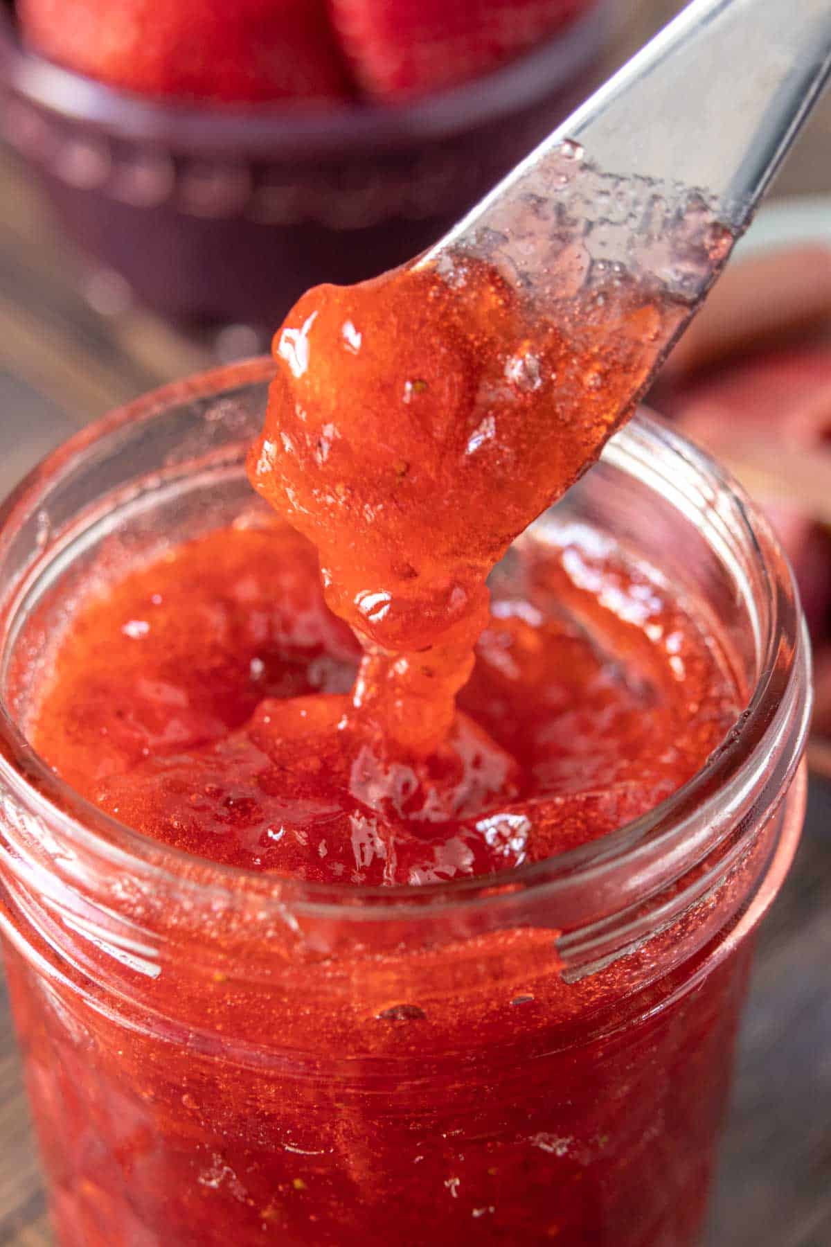 A spreader holding up a dollop of strawberry rhubarb jam just above the jam jar.