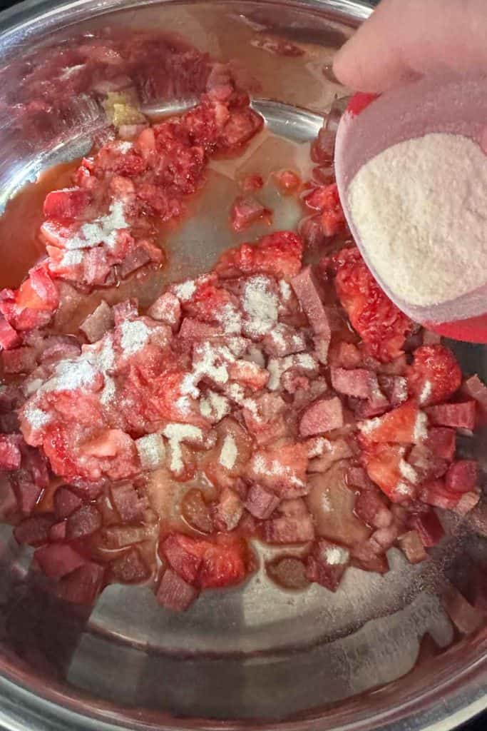 Powdered pectin being added to chopped rhubarb and mashed strawberries in a saucepan.