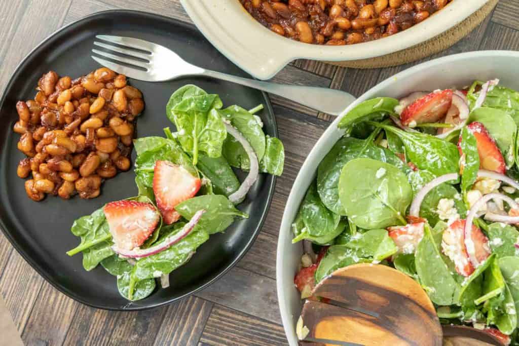 Overhead of strawberry spinach salad on a black plate with beans beside and serving bowl of salad.