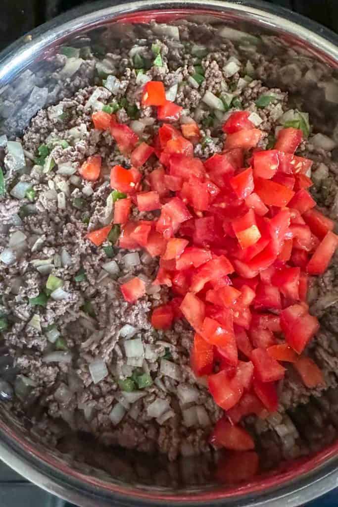 Diced tomatoes on top of cooked beef, onions, and peppers in a pan.