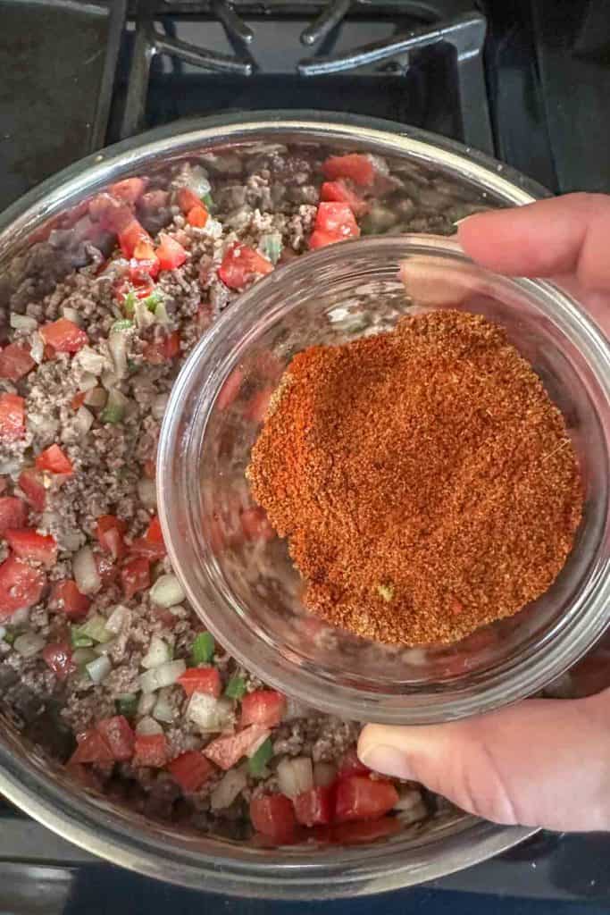 Spices in a small glass bowl held over a saute pan filled with beef and vegetables.