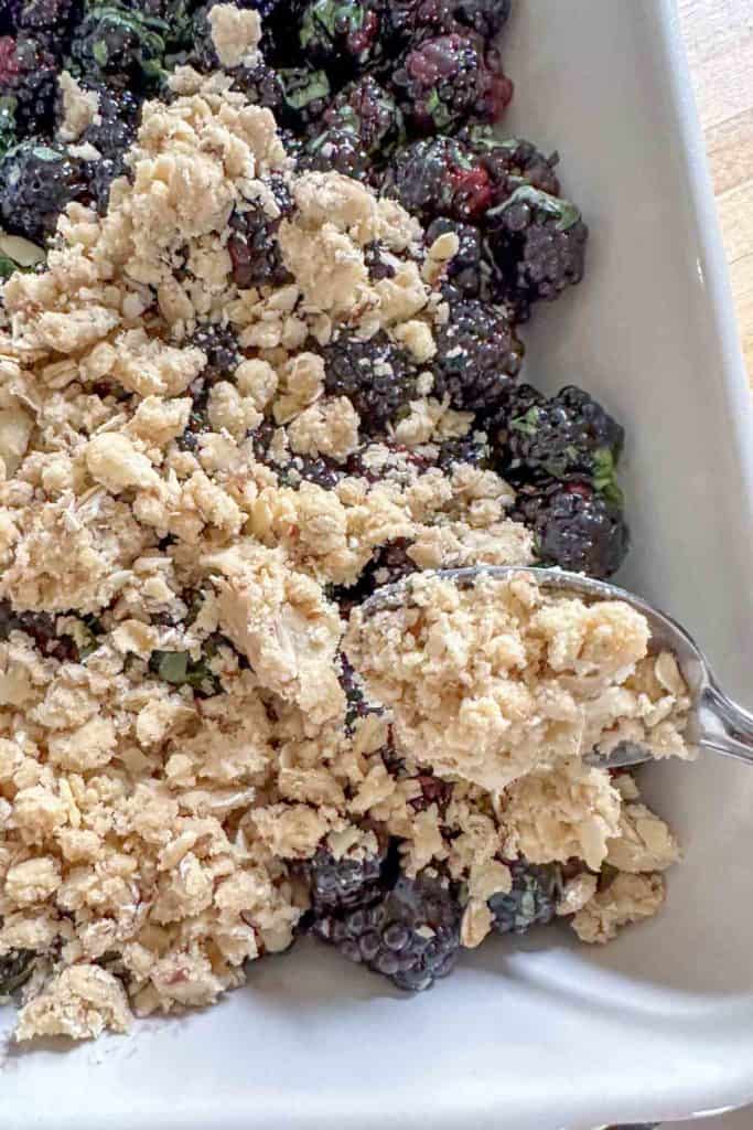 Scattering crisp topping over blackberries in a casserole dish.