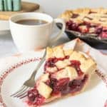 Slice of cherry pie on a white plate with red pattern, cup of coffee behind.