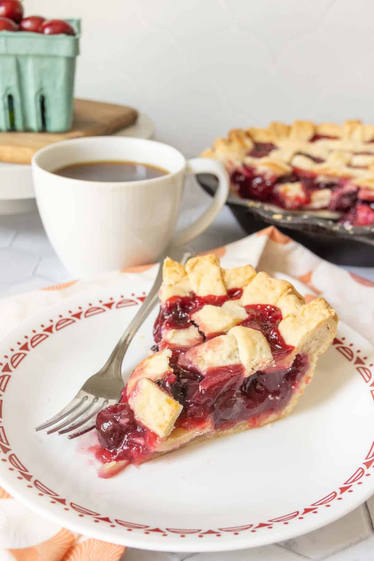 Slice of cherry pie on a white plate with red pattern, cup of coffee behind.