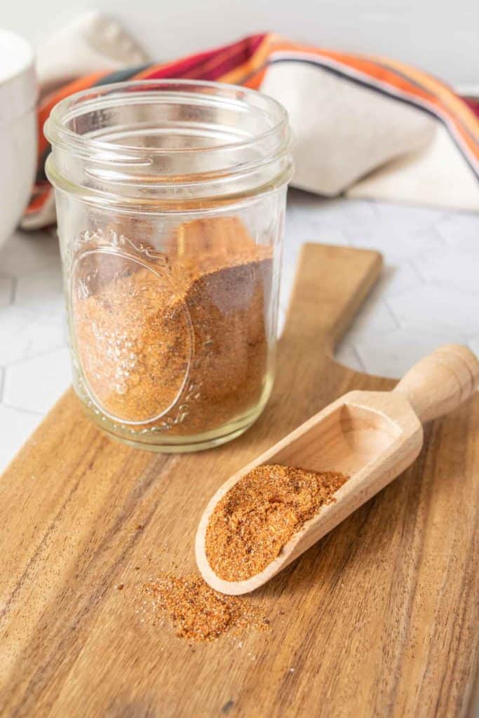 Jar of homemade taco seasoning next to a wooden scoop of seasoning on a cutting board.