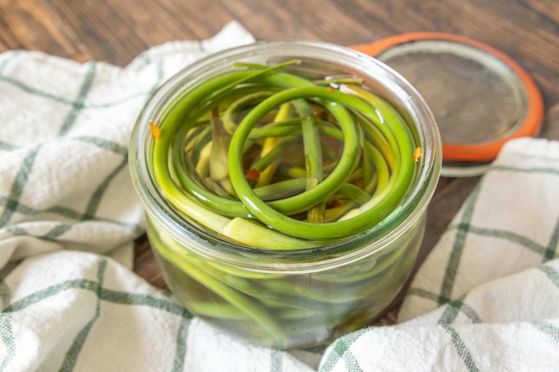 Open glass jar of pickled garlic scapes with a green and white napkin around it.