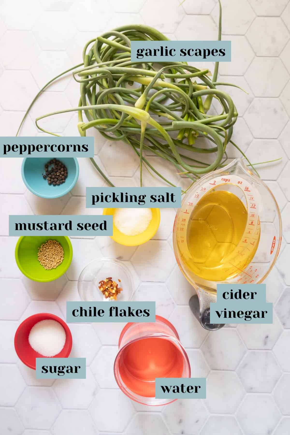 Ingredients for pickled garlic scapes on a tile surface with labels.