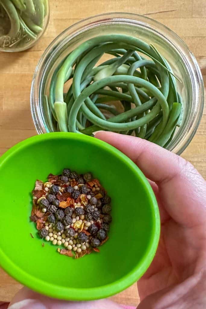 Bowl of spices in a green pinch bowl about to be added to a jar of garlic scapes.