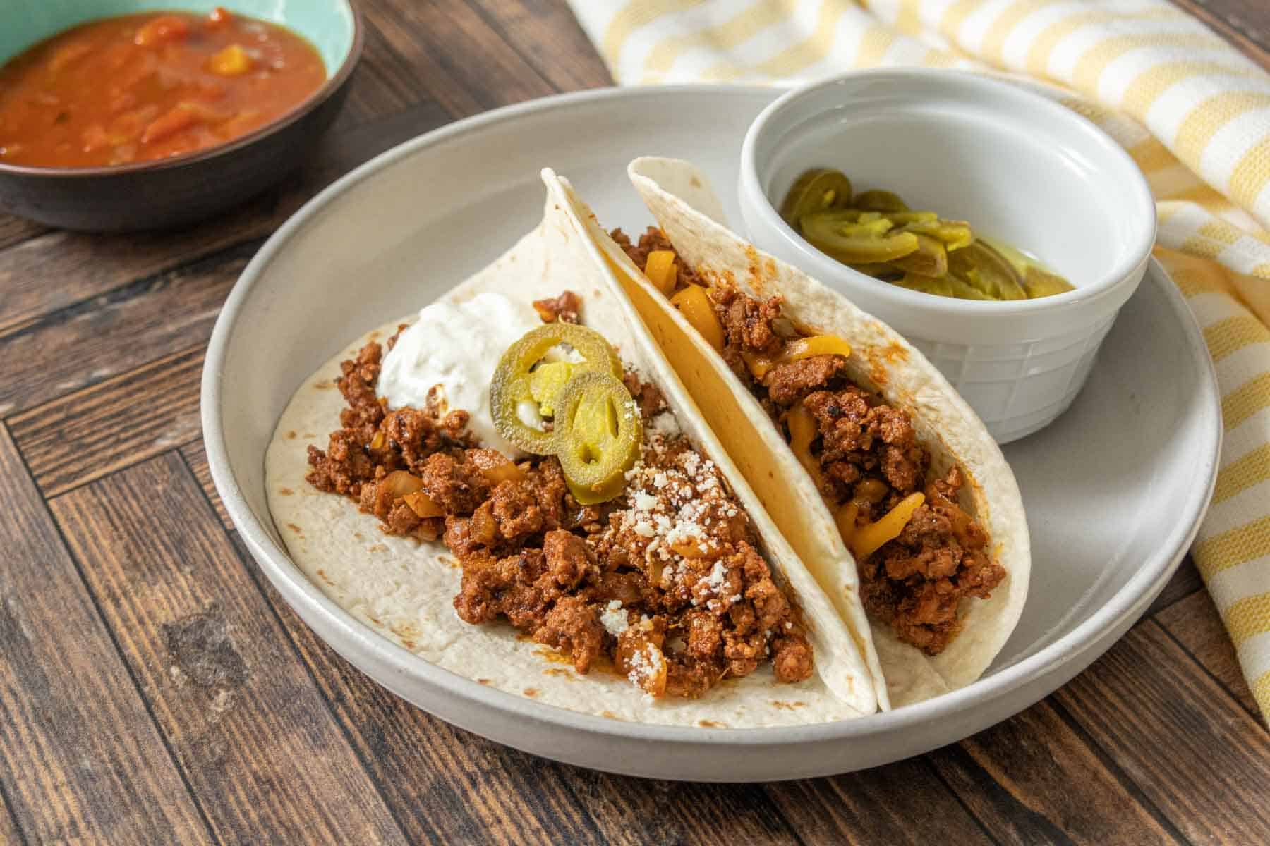 Two ground bison tacos on a plate with toppings and a bowl of pickled jalapenos.