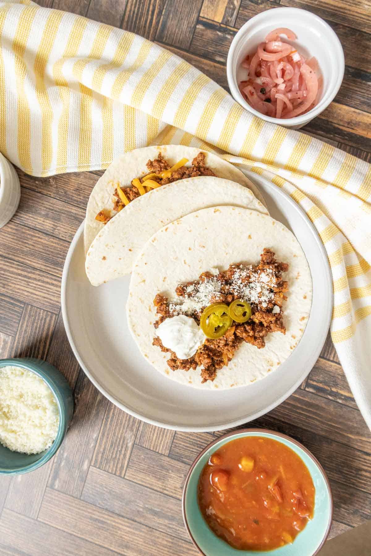 Overhead of ground bison tacos on a plate with toppings in bowls.