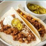 Two flavorful bison tacos in flour tortillas topped with cheese, sour cream, and jalapeños, served on a plate with extra sliced jalapeños on the side.