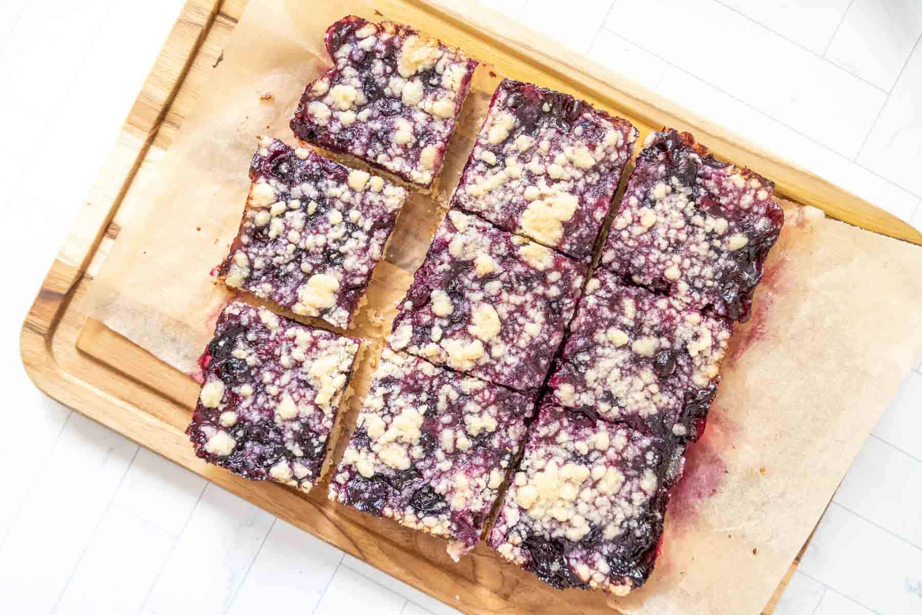 Overhead of sliced blueberry pie bars on parchment paper sitting on a cutting board.