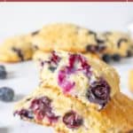 A close-up of blueberry scones stacked on a white surface, with juicy blueberries visible inside the scones and scattered nearby.