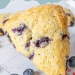 A close-up of a triangular blueberry scone with visible blueberries, next to fresh blueberries on a white surface. The text "BLUEBERRY SCONES" highlights this delicious display of homemade blueberry scones.