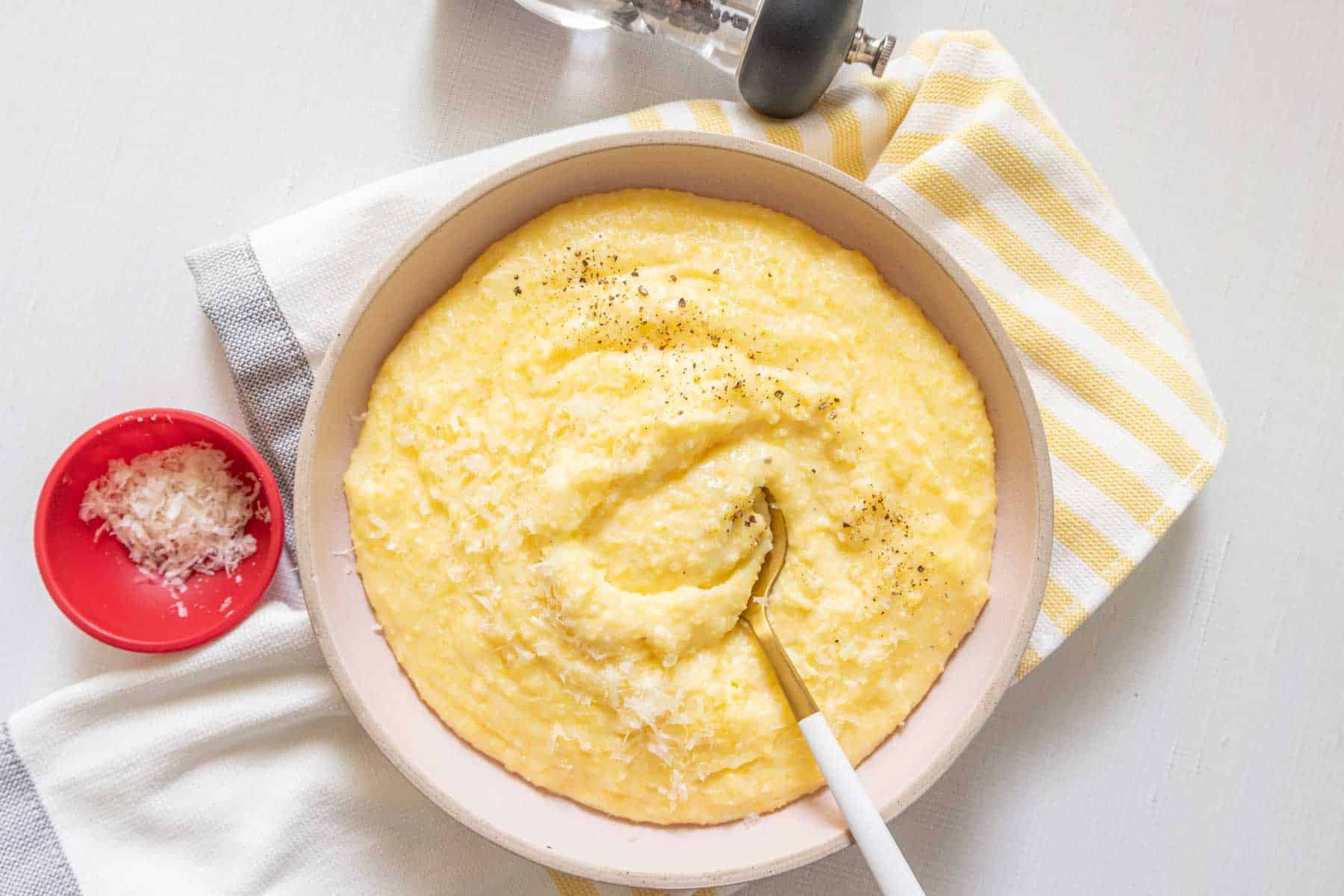 Overhead bowl of cheesy polenta with a spoon and a small red bowl of cheese nearby.