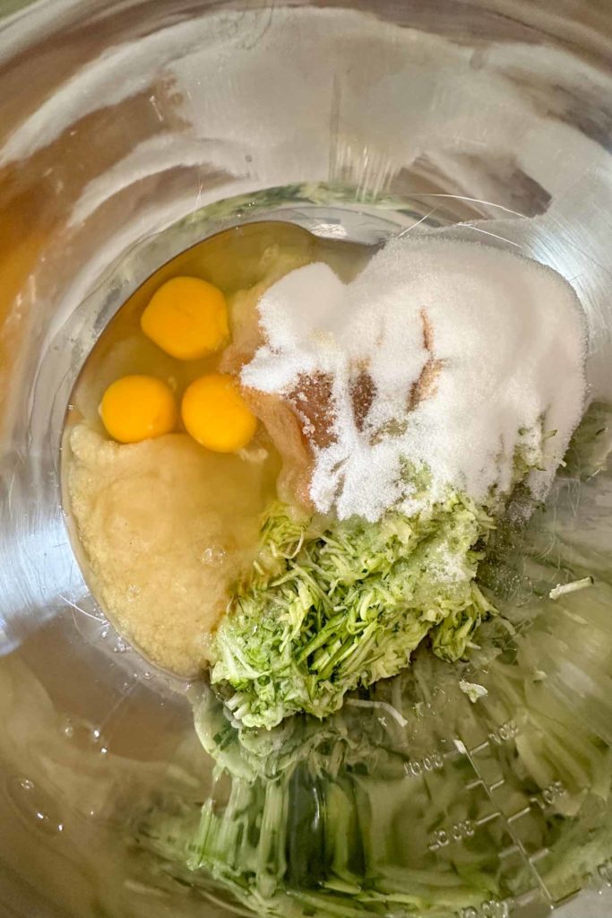 Shredded zucchini, sugar, eggs, applesauce, and vanilla in a metal mixing bowl.