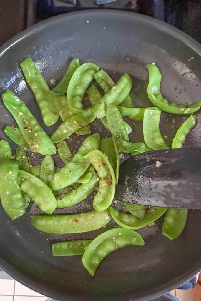 Stirring seasoning into cooked snow peas in frying pan.