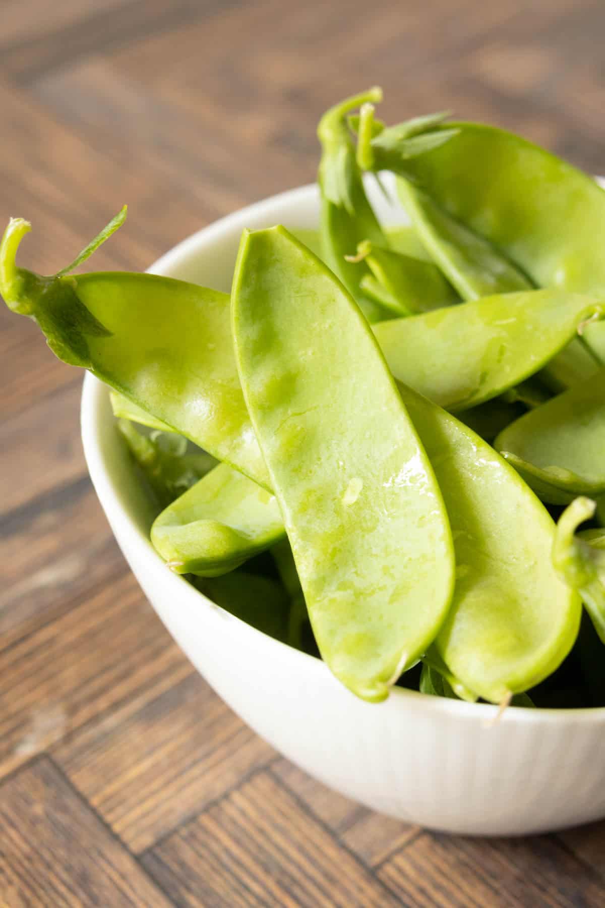 Raw snow peas in a white bowl on a wooden table.
