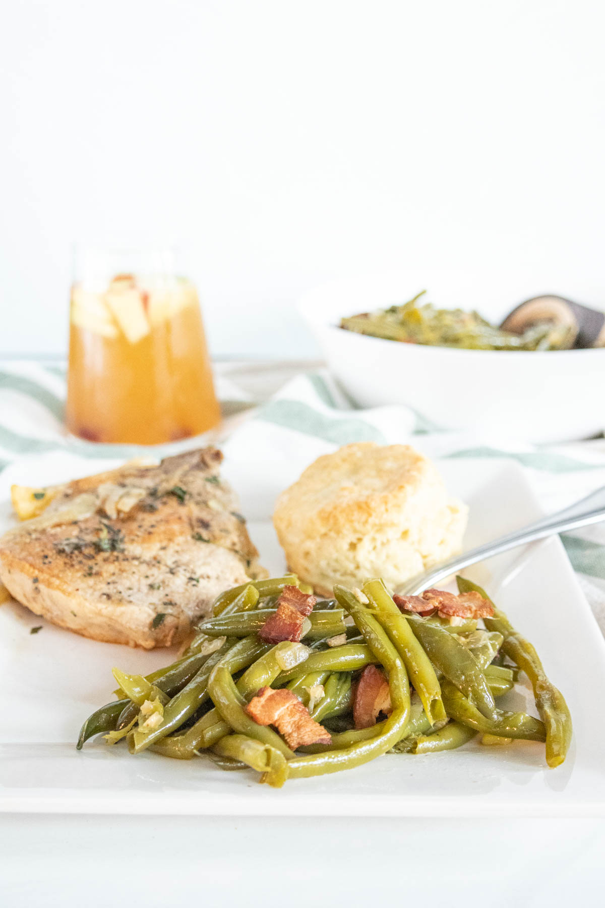 Dinner plate with southern green beans alongside a pork chop and biscuit with a glass of sangria in the background.