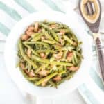 White bowl of Southern green beans on a green striped kitchen towel and a spoon beside.