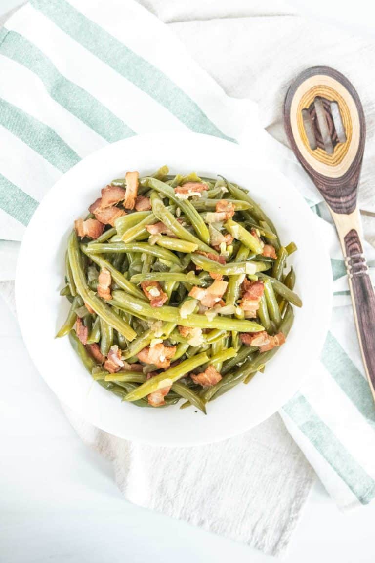 White bowl of Southern green beans on a green striped kitchen towel and a spoon beside.