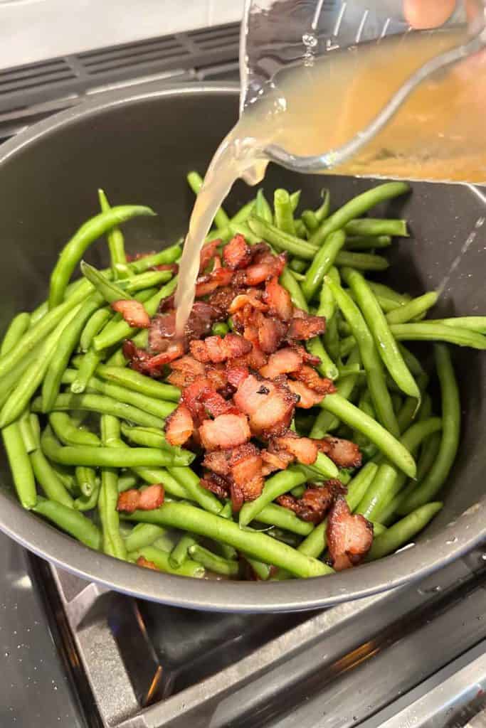 Pouring broth into a pot of green beans and bacon.