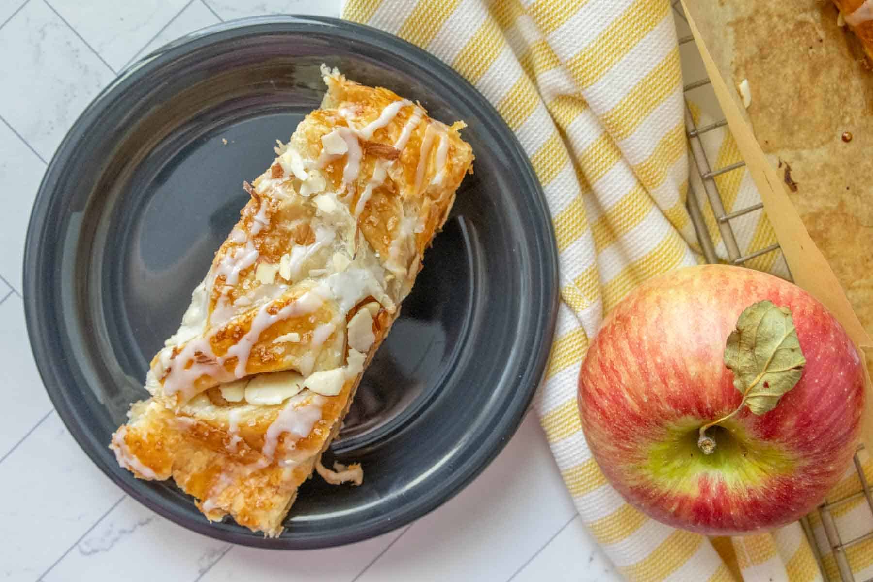 A slice of apple pastry on a plate next to an apple.